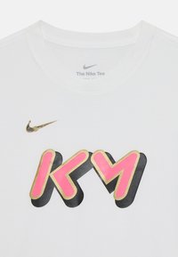 Biele tričko Nike s čiernou, ružovou a zlatou grafikou so snímkou písmen "KY." Zlaté logo Nike na ľavom ramene.
