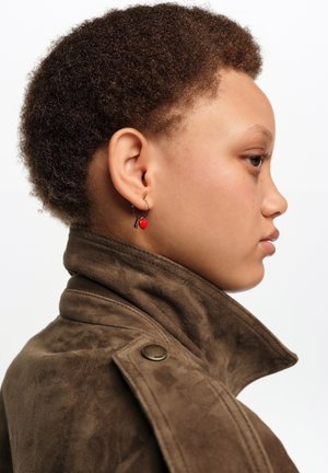 Młoda osoba o krótkich kręconych włosach, ubrana w brązową zamszową kurtkę, z kolczykiem-kołem ozdobionym czerwonym sercem, pokazana na białym tle z profilu.
