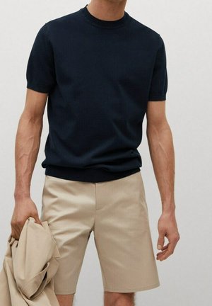 Mand iført en sort, kortærmet striktrøje og beige shorts, der holder en beige jakke i venstre hånd foran en ensfarvet baggrund.