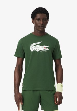 Hombre vistiendo una camiseta verde de Lacoste con un gran logo blanco de cocodrilo y pantalones deportivos verdes, sosteniendo una raqueta de tenis.
