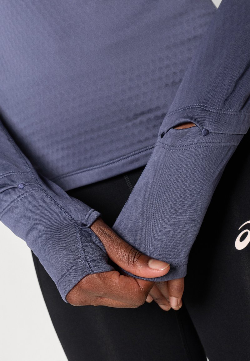 Long-sleeve athletic top in deep blue, featuring thumbholes, textured fabric, and flatlock seams. Paired with black high-waist leggings.