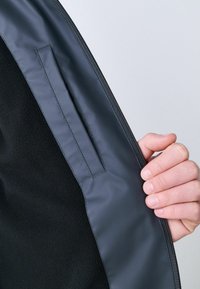 Crna fleeced jakna s glatkim tamnosivim unutarnjim slojem, koja ima zatvaranje na patentni zatvarač i vertikalnu džepu na unutrašnjoj strani.