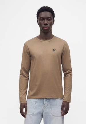 Junger Mann trägt ein braunes Langarmshirt mit kleinem Logo auf der Brust und hellblaue Jeans, steht vor einem schlichten weißen Hintergrund.