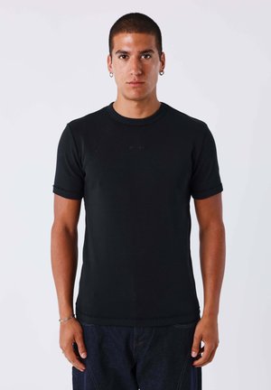 Homme aux cheveux courts et foncés et boucles d’oreilles, debout face à l’avant, portant un t-shirt noir ajusté à manches courtes et un jean foncé sur fond uni.