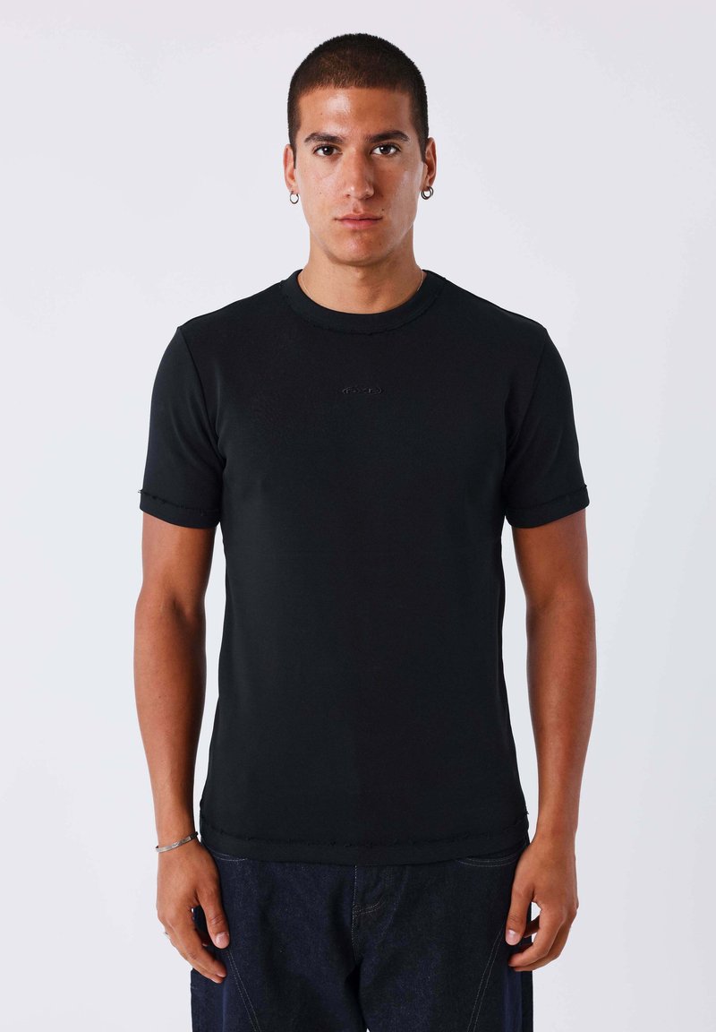 Homme aux cheveux courts et foncés et boucles d’oreilles, debout face à l’avant, portant un t-shirt noir ajusté à manches courtes et un jean foncé sur fond uni.