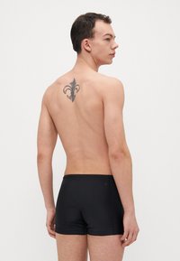 Juodos maudymosi šortai, prigludęs dizainas, lygi tekstūra, minimalistiškas stilius, dėvimi modelio su tatuiruote ant nugaros.