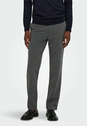 Homme portant un pantalon de costume gris, des mocassins noirs et une chemise à manches longues foncée, debout devant un fond clair uni.