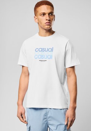 Młody mężczyzna o kręconych rudych włosach, ubrany w białą koszulkę z napisem "casual" w jasnoniebieskim kolorze oraz jasnoniebieskie szorty typu cargo, stoi na szarym tle.