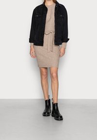 Veste en denim noir portée sur une robe beige en maille côtelée avec une ceinture nouée à la taille. Bottines noires avec un détail de languette sur le talon.