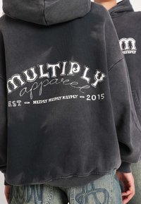 Sweat à capuche gris foncé, avec un grand texte blanc "MULTIPLY apparel" et une finition usée. Le texte inclut l'année d'établissement 2015.