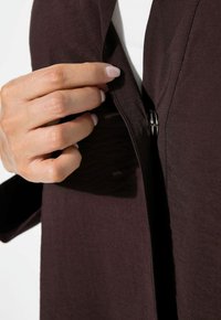 Bruine, lichte kleding met een gestructureerde afwerking. Een hand opent de rits, wat een strak ontwerp en schone lijnen laat zien.