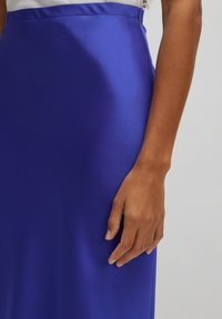 Satijnen blauwe rok met een gladde, glanzende textuur. Heeft een aansluitende taille en een rits aan de zijkant. Close-up van een hand die op de stof rust.