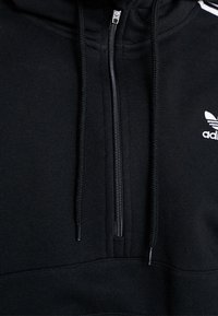 Svart zip-up-hoodie i mjukt tyg med hög krage, med vita detaljer och Adidas-logotypen på övre vänstra sidan.