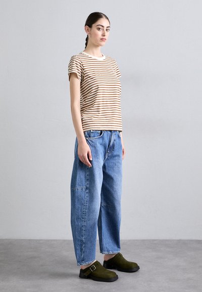 Riba pruun ja beež triibuline puuvillane T-särk, lõdvad sinised denim püksid ja rohelised sametkingad klambriga. Mudel poseerib neutraalse tausta ees.