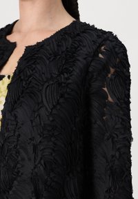 Black textured jacket with layered fabric, featuring a floral pattern, rounded collar, and sheer accents at the sleeves.