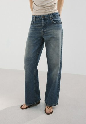 Persoon die een losse blauwe jeans met hoge taille en een beige top draagt, blootsvoets staand op een lichte vloer tegen een effen achtergrond.