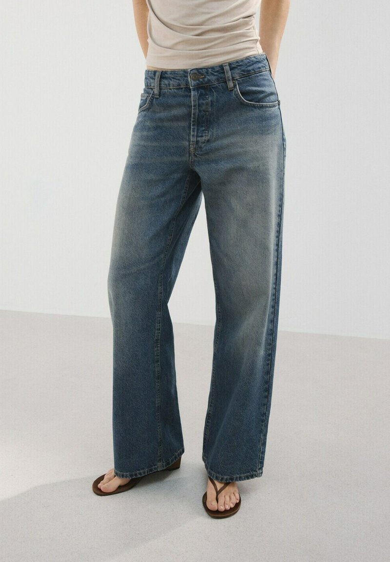 Person trägt hoch taillierte, lockere blaue Jeans und ein beiges Oberteil, steht barfuß auf einem hellen Boden vor einem schlichten Hintergrund.