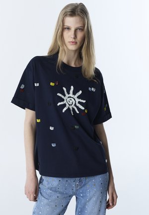 Joven con cabello rubio largo que lleva una camiseta azul marino con un diseño de sol blanco y pequeñas formas bordadas de colores, combinada con vaqueros de color azul claro.