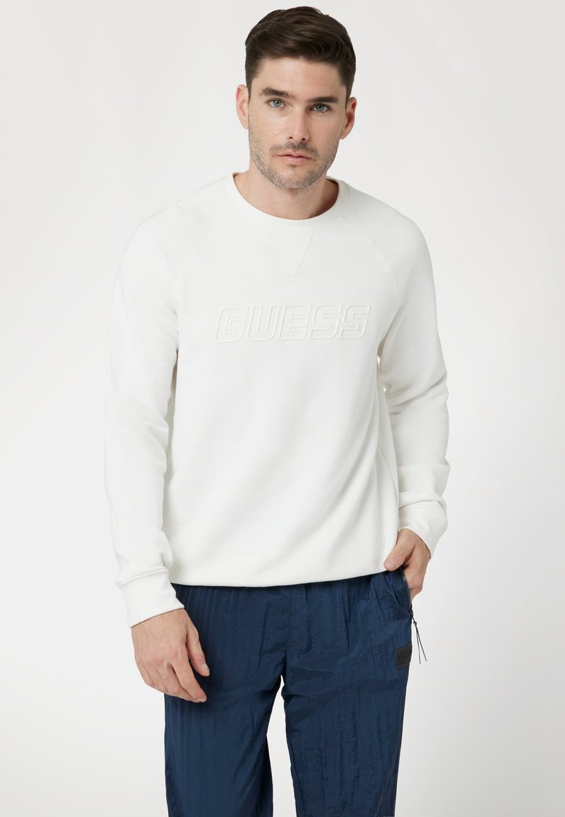 Felpa bianca con logo "GUESS" in rilievo, polsini a coste e scollatura a girocollo. Abbinata a pantaloni blu navy strutturati.