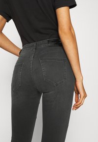 Jeans de color gris oscuro con un diseño ajustado, estilo de cinco bolsillos y costuras en contraste. Presenta una etiqueta texturizada en la cinturilla y un ligero estiramiento.