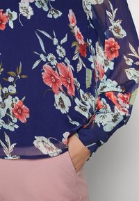 Blouse fleurie avec fond bleu marine, présentant des fleurs rouges et bleu clair. Manches longues et transparentes avec boutons en accent. Associée à un pantalon rose.