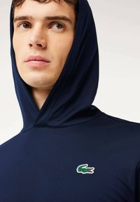 Marinblå huvtröja i mjukt tyg med en liten grön och vit logotyp på bröstet. Närbild av halsringning och huva.