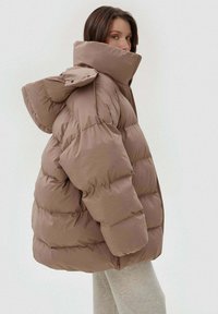 Brązowa ocieplana kurtka w stylu oversize z wysokim kołnierzem i kapturem. Ma pikowane sekcje oraz gładką, błyszczącą powierzchnię.