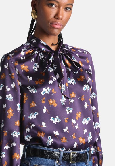 Femme aux cheveux tressés portant des boucles d'oreilles en or et un chemisier violet fleuri avec un nœud au cou, rentré dans un jean avec une ceinture noire.