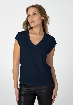 Woman with light brown hair wears a navy blue cap sleeve V-neck top and black leather pants, looking slightly to her right.