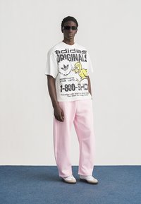 Fiatal férfi napszemüvegben, fehér, grafikus Adidas Originals pólóban, bő rózsaszín nadrágban és fehér sportcipőben áll a kék padlón.