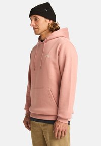 Pink hooded sweatshirt made of soft fabric, featuring a kangaroo pocket, drawstrings, and a small logo on the chest. Worn with a black beanie.
