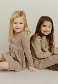 Dvije djevojke sjede jedna pored druge, obučene u svijetlosmeđe pletene haljine. Haljine imaju duge rukave, slojeviti dizajn i suptilnu cvjetnu vezu.