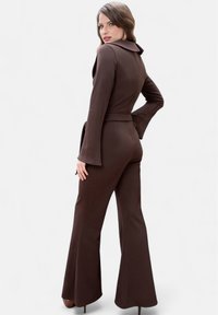 Hnědý upnutý overal s límečkem, dlouhými rozšířenými rukávy a pásem v oblasti pasu. Vyrobený z hladkého materiálu, design obsahuje zip na zádech.