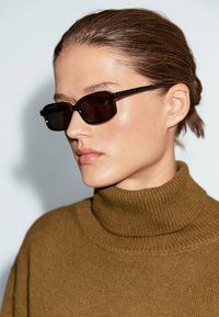 Occhiali da sole neri rettangolari con lenti scure, indossati con un maglione a collo alto in cotone marrone. Texture liscia con minimi dettagli.