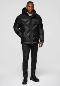 Giacca imbottita nera con design trapuntato, dotata di cappuccio, chiusura con zip e tasche laterali, indossata sopra pantaloni neri e stivali.