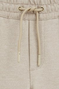 Bežīgas bikses ar teksturētu audumu, ar šņorējamu vidukli un plastikāta uzgaļiem. Redzamas šuvju detaļas ap kabatām.