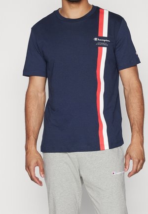 Mann trägt ein marineblaues Champion-T-Shirt mit roten und weißen vertikalen Streifen und graue Champion-Sweathosen, steht vor einem einfarbigen Hintergrund.