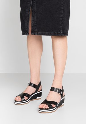 Jambes portant des sandales compensées noires avec des brides à la cheville et des boucles argentées, associées à une jupe noire longueur genou avec une fente devant.