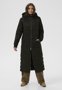 Dlhý čierny prešívaný kabát so zapínaním na zips, kapucňou a bočnými vreckami. Obsahuje panelové švy a patentky na leme. Nosený nad hnedými nohavicami.