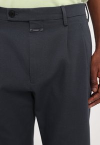 Gros plan d'une personne portant un pantalon gris foncé sur mesure avec une taille boutonnée et une petite étiquette "CLOSED" au-dessus de la braguette.