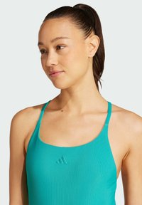 Tyrkysový žebrovaný tílko s tenkými ramínky a vyšitým logem na přední straně, které se vyznačuje přiléhavým střihem a jemnou texturou.