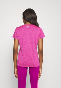 Mujer con cabello largo y oscuro, vistiendo una camiseta rosa de manga corta y leggings morados brillantes, de espaldas sobre un fondo gris claro liso.