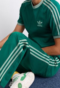 Grön atletisk träningsdräkt med matchande överdel och byxor, med tre vita ränder på axlarna och benen, samt en broderad logotyp.