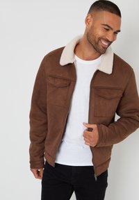 Brun faux suede jakke med en beige shearling krave, front lynlås, to brystlommer og en glat tekstur, parret med en hvid t-shirt.