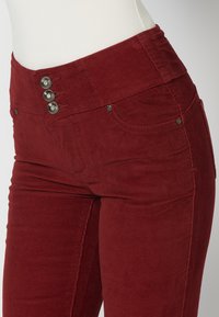 Červené manšestrové kalhoty s vysokým pasem, třemi kovovými knoflíky a předními kapsami, s vertikálním ribbovaným vzorem a slim fit siluetou.
