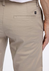 Beige puuvillahousut, joissa on sileä pinta, takatasku ja pieni musta logo-täg oikealla puolella.