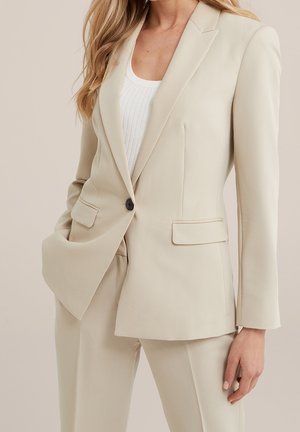 Kvinde iført beige skræddersyet blazer og matchende bukser over en hvid ribbet top, poserer med den ene hånd i lommen.