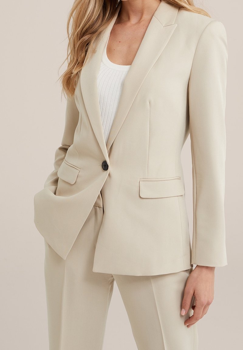Femme portant un blazer beige cintré et un pantalon assorti sur un top blanc côtelé, posant avec une main dans la poche.