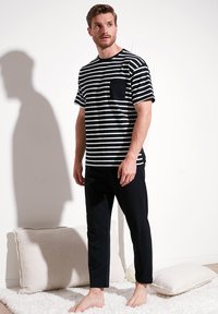 Tricou cu dungi alb-negru, cu mâneci scurte și buzunar, combinat cu pantaloni negri sportivi. Modelul stă pe un covor deschis.