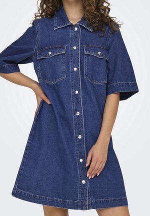 Vrouw draagt een blauw denim overhemdjurk met korte mouwen en twee borstzakken, poseert met één hand op de heup.